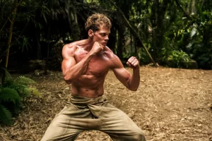 Muscular man with no shirt performing a martial arts stance in a forest.