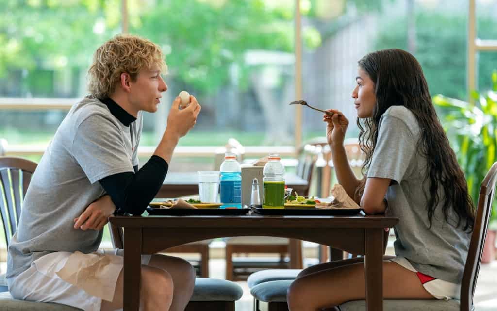 Two young adults eating and conversing at a cafeteria table, with drinks and salads in front of them.