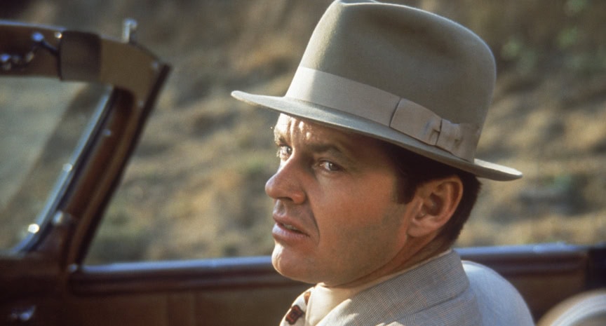Man in a vintage suit and fedora hat sitting in a car, looking away from the camera with a serious expression.