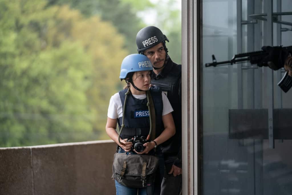 A journalist wearing a "press" helmet and vest holds a camera, looking apprehensive, as an armed person in tactical gear stands behind her, both gazing out of a window.