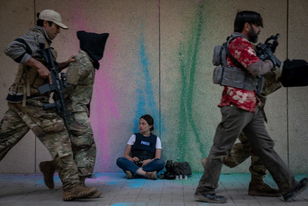 A seated individual labeled 'press' observing armed personnel in motion against a backdrop of a colorful wall.