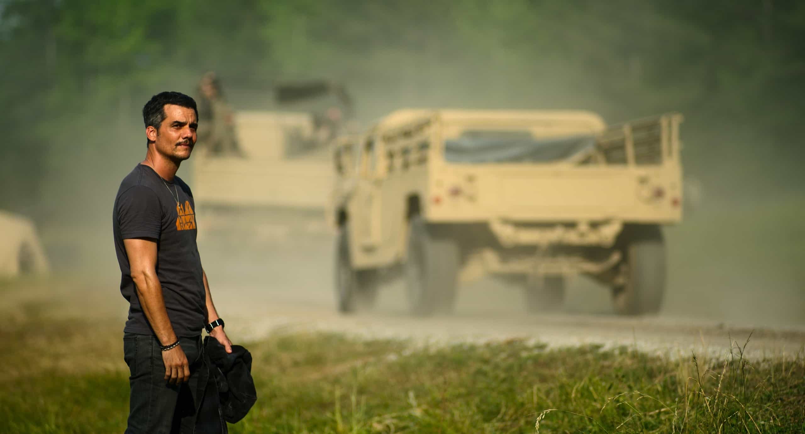Man standing by the roadside with military vehicles passing by in a dusty environment.