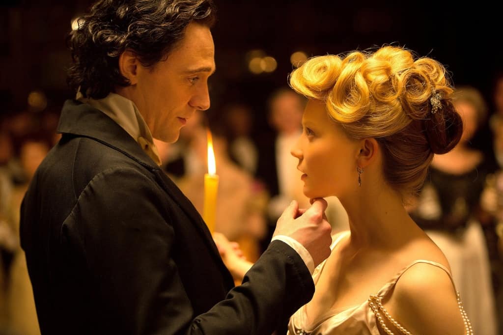 A man in a black coat gently touches a woman's chin while she, wearing a golden gown with an elaborate updo, looks at him intently in a candlelit room.