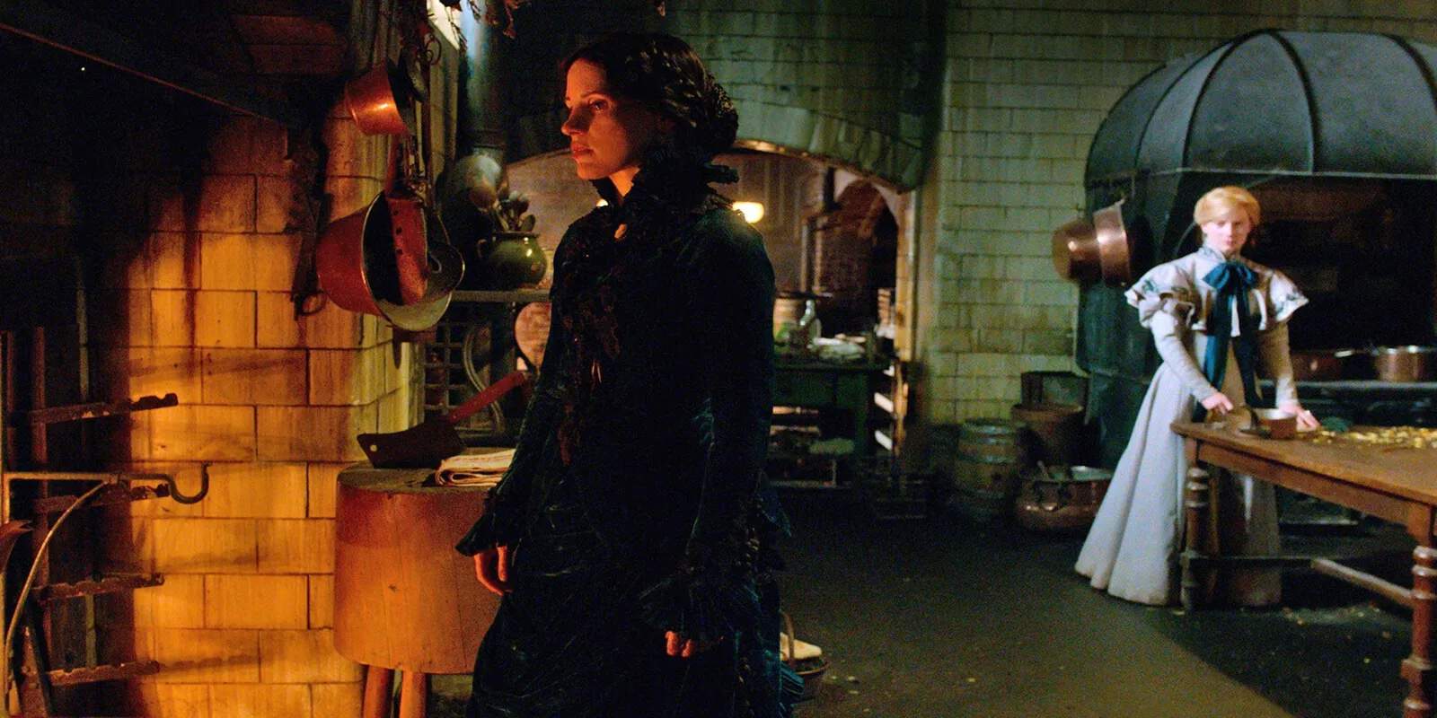 A woman in a dark feathered dress stands in a grim, dimly-lit kitchen, facing another woman in a pale blue medieval gown who is standing by a large oven.