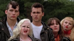 A group of five individuals styled in 1950s fashion, featuring leather jackets and bold hairstyles, displaying serious expressions in an outdoor setting.