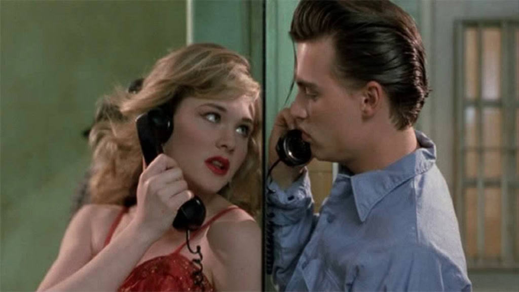 A young woman in a red dress talking on an old-fashioned telephone, looking surprised at a young man in a blue shirt who is close to her, both inside a room with green walls.