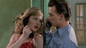 ‘Cry-Baby’ 4K UHD Blu-Ray Review – John Waters Classic Gets A 5-Star Presentation A young woman in a red dress talking on an old-fashioned telephone, looking surprised at a young man in a blue shirt who is close to her, both inside a room with green walls.