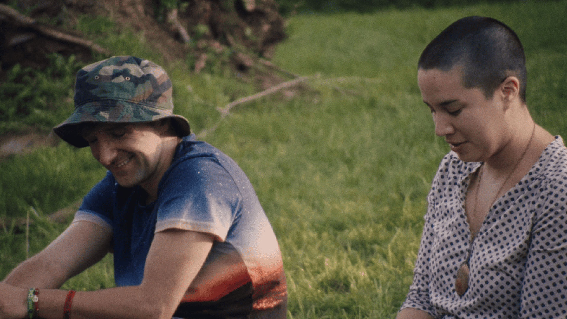 A man in a camouflage hat and a woman with a shaved head sitting outside, engaged in a casual conversation near greenery.