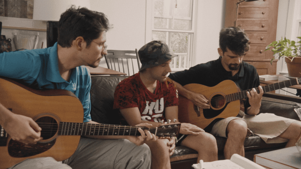 Three men sitting on a couch, playing acoustic guitars and looking at sheet music in a cozy living room.