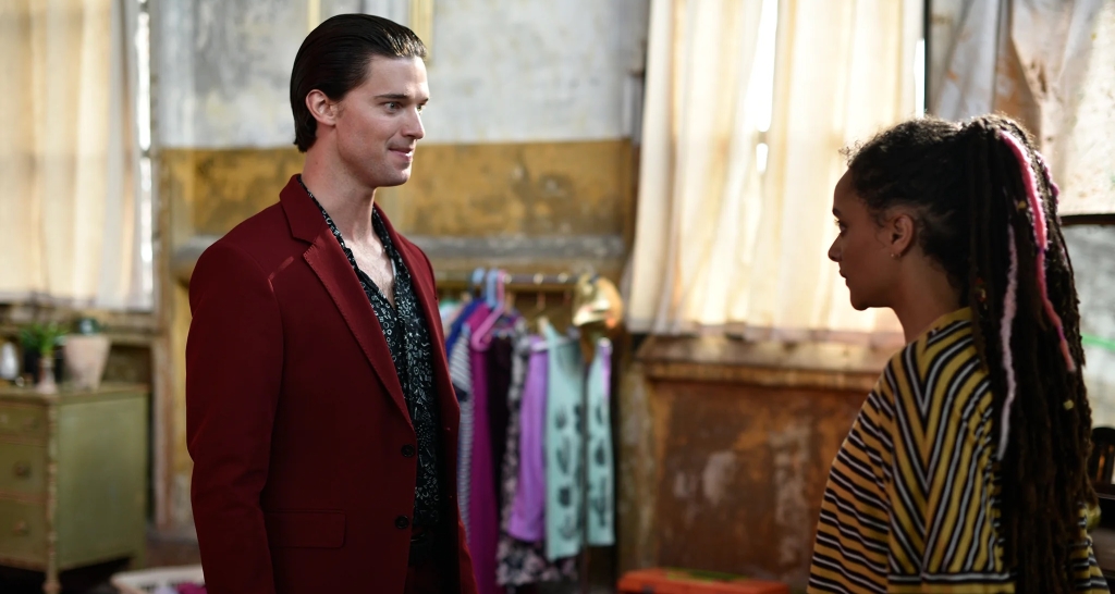 A man in a red blazer smiling at a woman with braided hair in a striped top, standing in a room with a rustic interior.