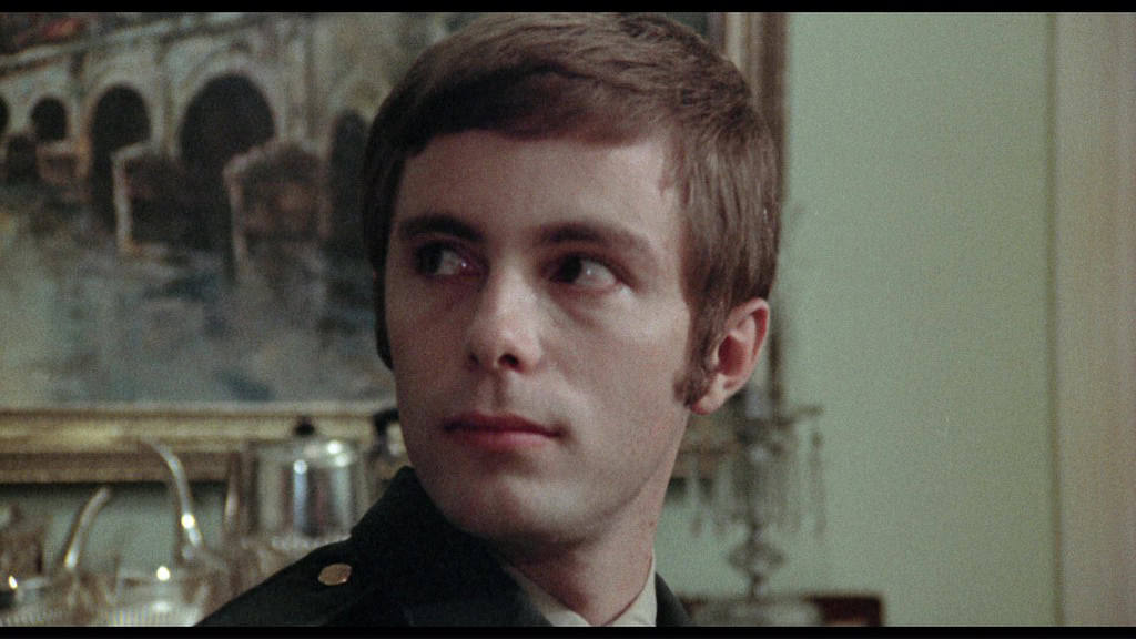 Young man in a military uniform, with short dark hair and a solemn expression, sitting indoors with a classical painting in the background.