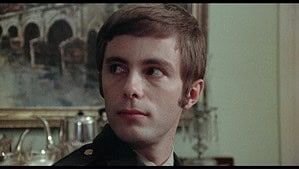 Young man in a military uniform, with short dark hair and a solemn expression, sitting indoors with a classical painting in the background.