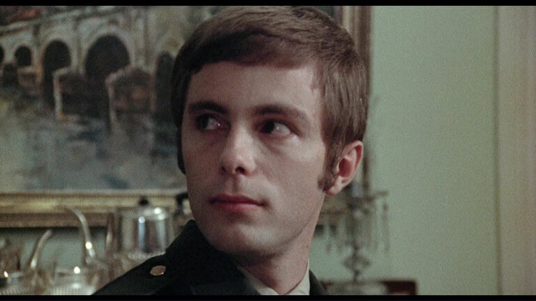 Young man in a military uniform, with short dark hair and a solemn expression, sitting indoors with a classical painting in the background.