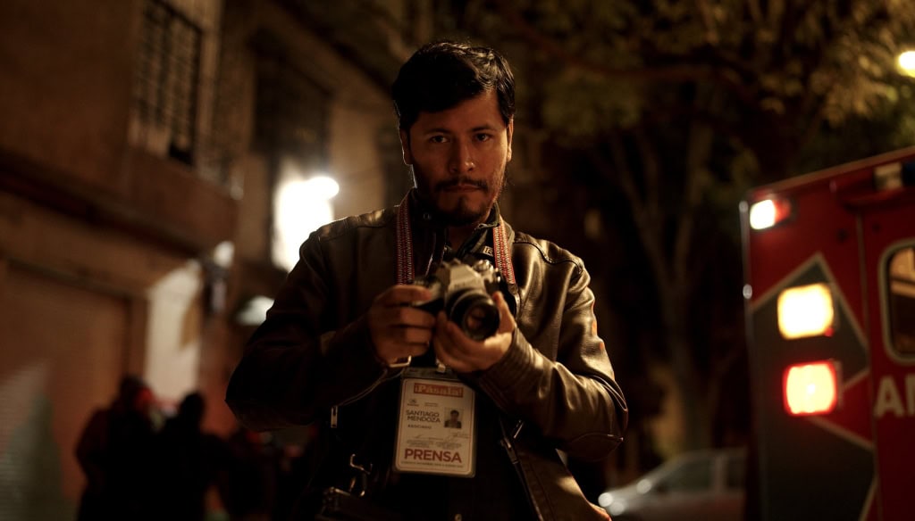 A male photojournalist with a press pass, holding a camera, stands on a dimly lit street at night with an ambulance in the background.