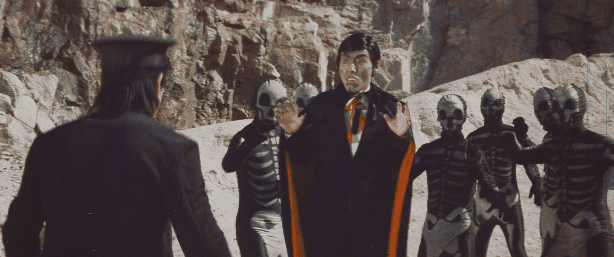 A person in a cape and costume surrounded by individuals in skeletal outfits in a rocky setting.