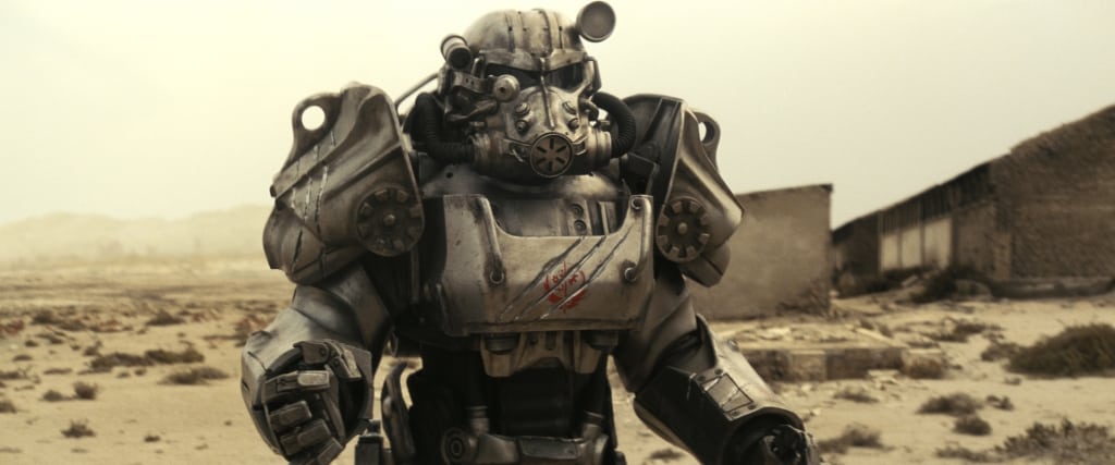 A mechanized suit or robot standing in a desolate, sandy landscape with abandoned structures in the background.