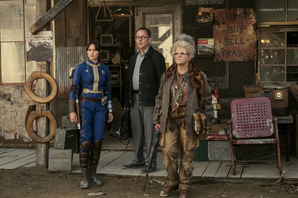 Three characters from a film, dressed in distinct costumes, stand in a rustic, cluttered shop interior looking intently to their left.