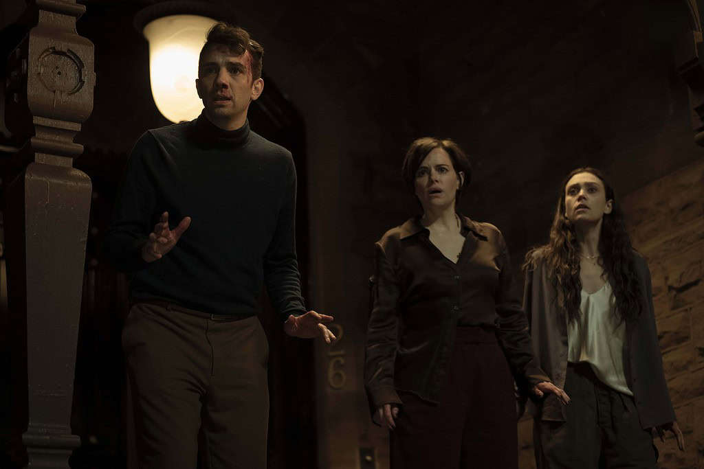 Three people look anxious in a dimly lit room, with an old clock in the background showing 12:06.
