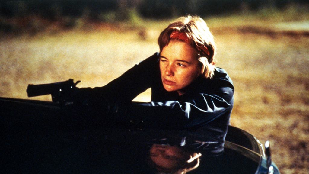 A person wearing a black jacket and holding a gun leans out of a car window with a focused expression.