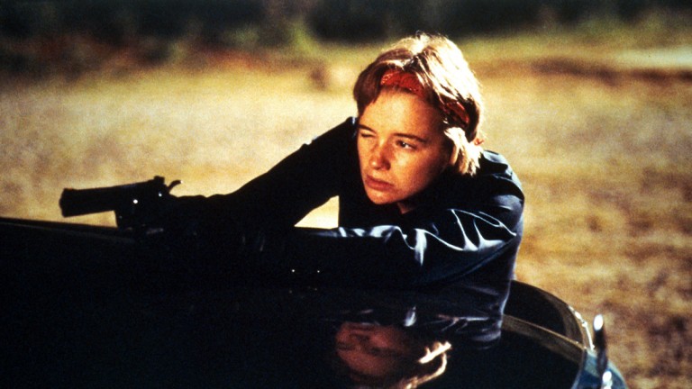 A person wearing a black jacket and holding a gun leans out of a car window with a focused expression.