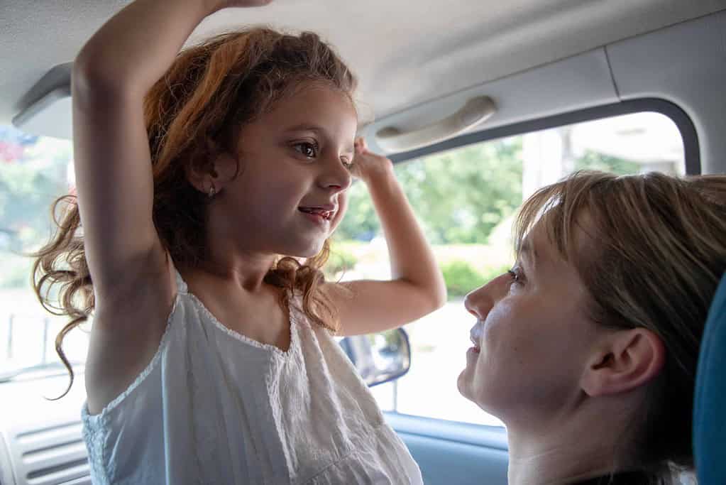 A young girl standing in a car raises her arm, smiling at a woman seated beside her who is looking up at her with affection.