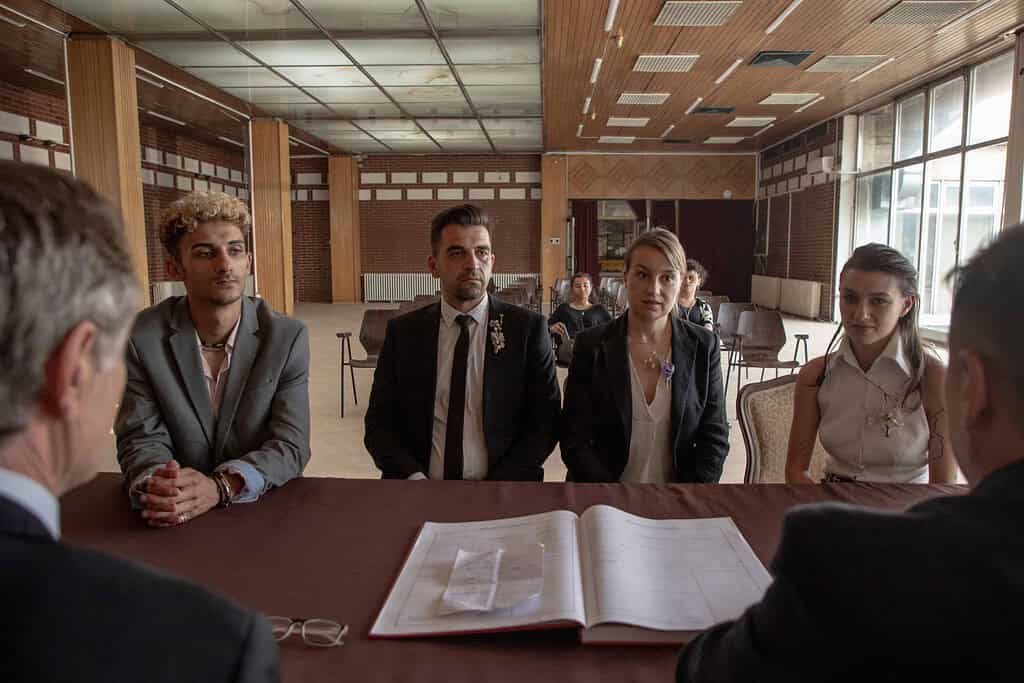 Five individuals engaged in a serious meeting at a table with documents visible; they look attentively towards a person speaking out of frame.