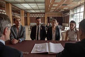 Five individuals engaged in a serious meeting at a table with documents visible; they look attentively towards a person speaking out of frame.