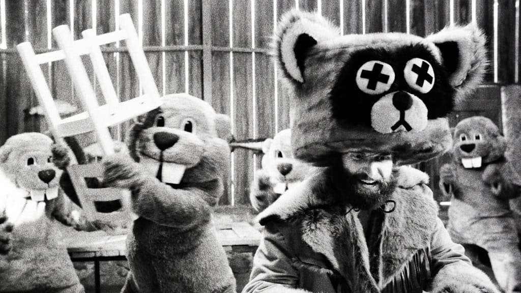 Black and white image depicting a man in a bear costume surrounded by others in similar costumes, holding objects, in a rustic setting.