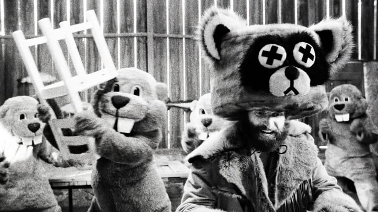 Black and white image depicting a man in a bear costume surrounded by others in similar costumes, holding objects, in a rustic setting.