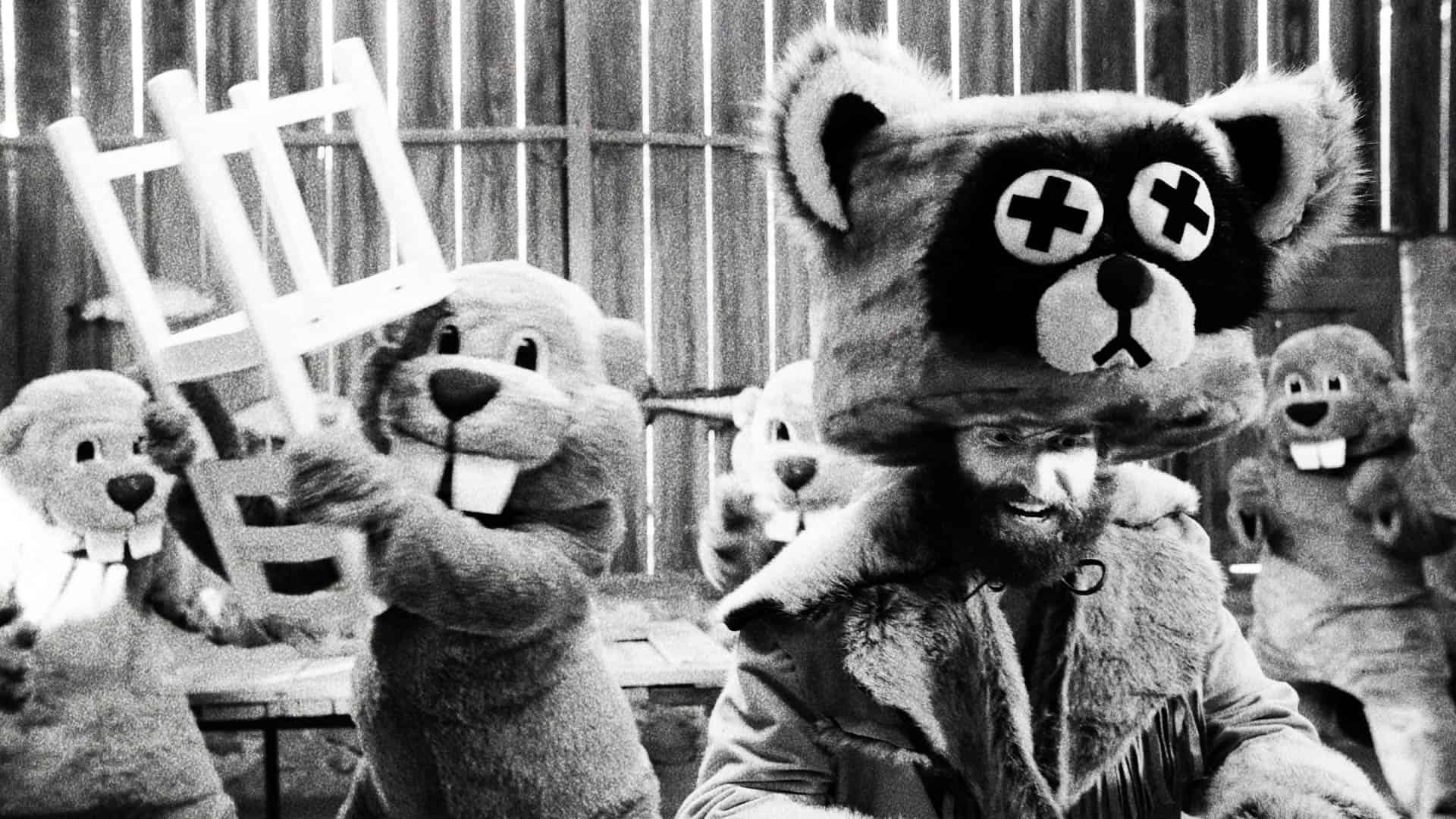 Black and white image depicting a man in a bear costume surrounded by others in similar costumes, holding objects, in a rustic setting.