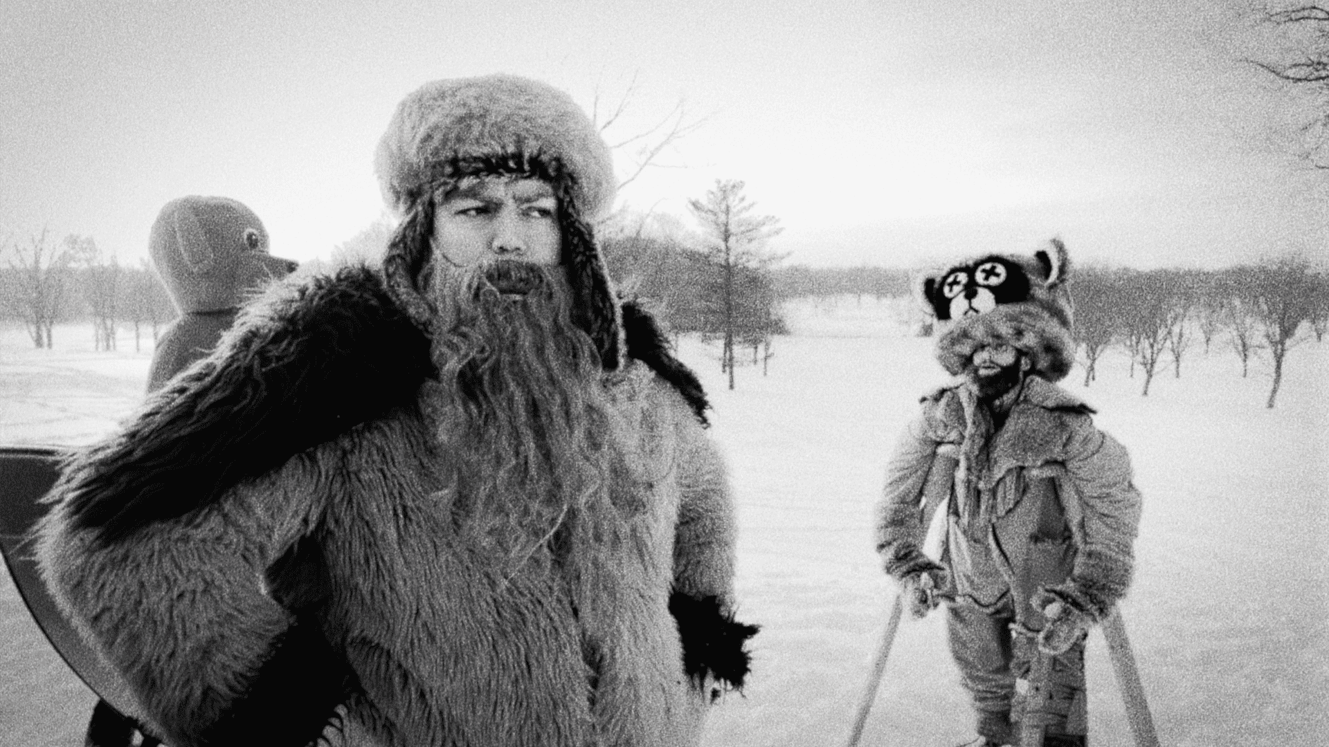 A black and white photo of two people in furry winter costumes, one dressed as a bear and another as a raccoon, standing in a snowy landscape.
