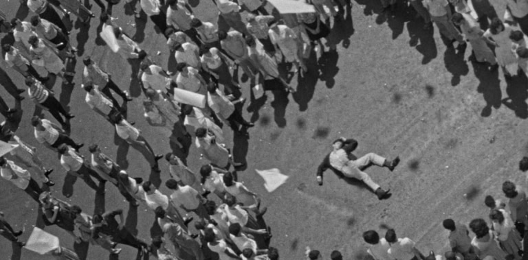 Aerial view of a single person lying on the ground surrounded by a crowd of protesters, some holding signs.