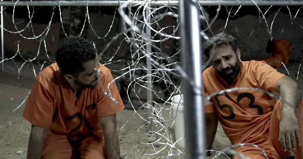 Two men in orange jumpsuits sitting behind barbed wire, appearing distressed and conversing in a dimly lit confinement area.