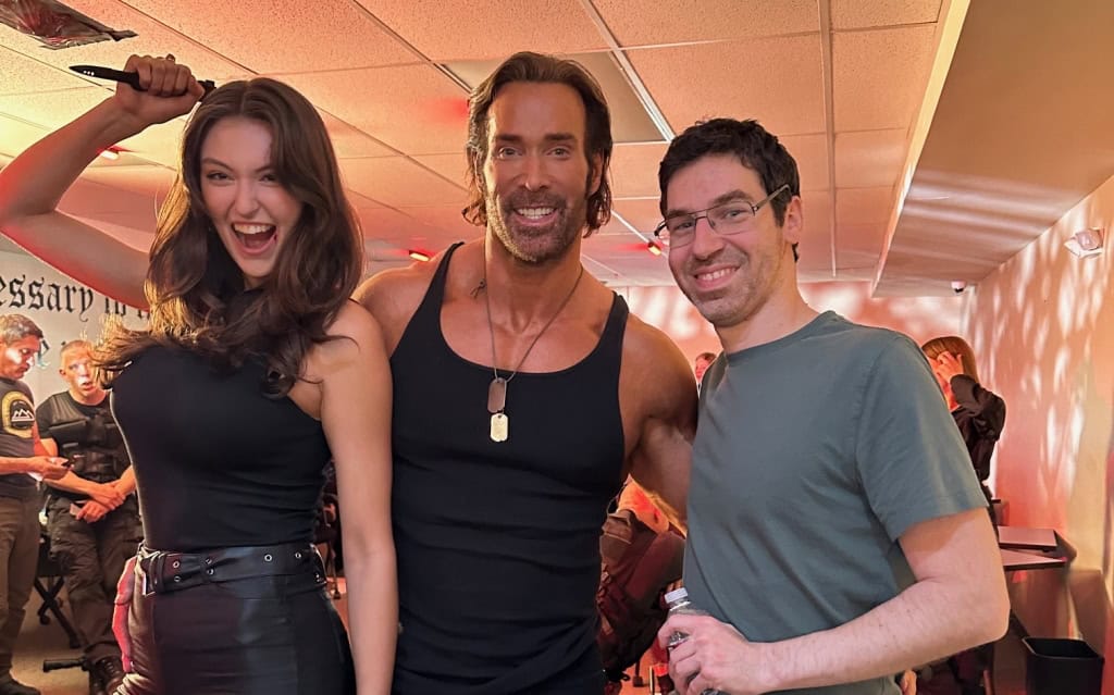 A woman and two men posing, the woman holding a sword above her head, set in a room with other people and equipment in the background.