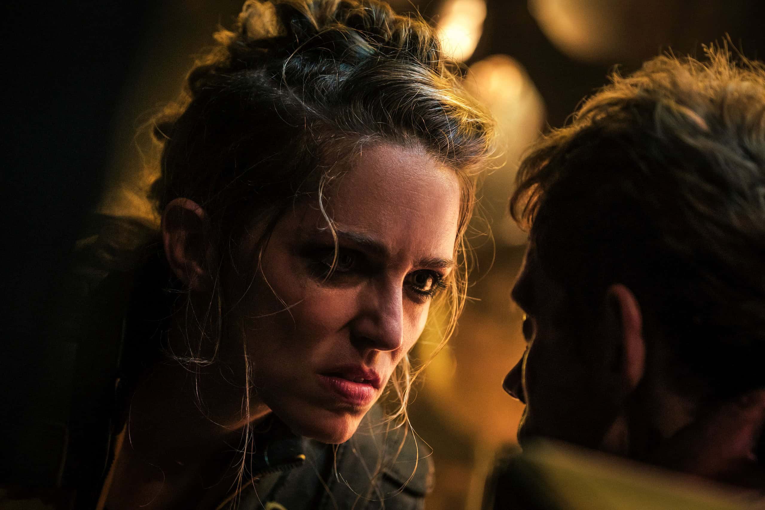 A close-up of a woman intensely staring at a man, showing concern or confrontation, with dramatic lighting highlighting their faces.
