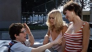 ‘Joysticks’ Blu-Ray Review – Hitting The Hallmarks Of ’80s Sex Comedies A man in glasses sitting on a car hood converses with two smiling women standing beside him in a sunny parking lot.