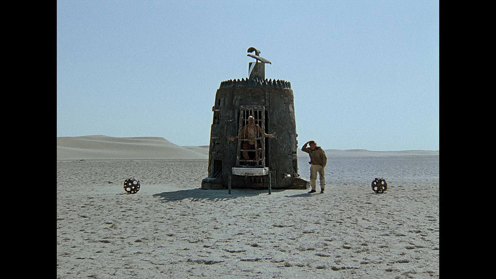 A wooden hut on a desert landscape with a person on top and two others beside it, under a clear sky.