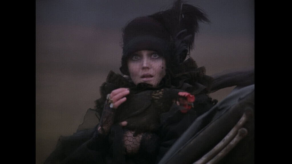 A woman in elaborate dark victorian attire and makeup looks startled, with large expressive eyes, holding a feathered hat.