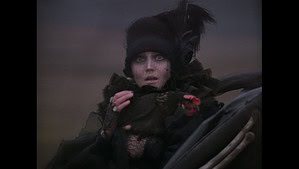 ‘The Savage Hunt Of King Stakh’ Deluxe Edition Blu-Ray Review – An Eerie & Surreal Folk Horror Gem A woman in elaborate dark victorian attire and makeup looks startled, with large expressive eyes, holding a feathered hat.