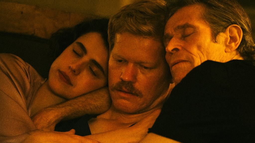 Three people, two men and one woman, emotionally embracing, resting heads together with somber expressions in a dimly lit room.