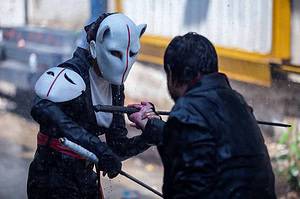 Two individuals in a dramatic outdoor scene, one wearing a white and black mask, engaging in a staged sword fight.