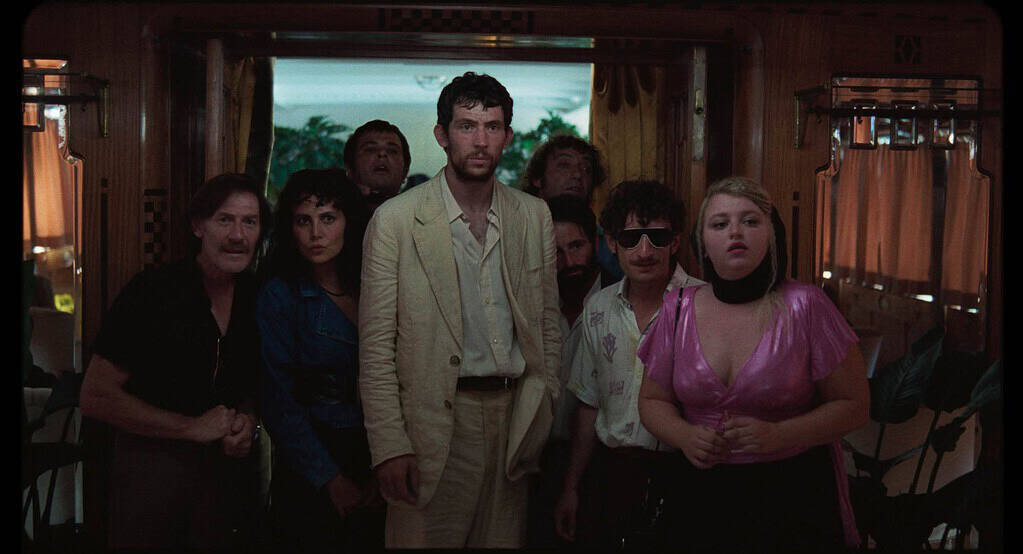 A group of eight diverse people stand in a dimly lit vintage trailer, looking intently at the camera with serious expressions.