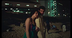 A young man and woman walking closely together on a beach at night, with an industrial structure in the background.