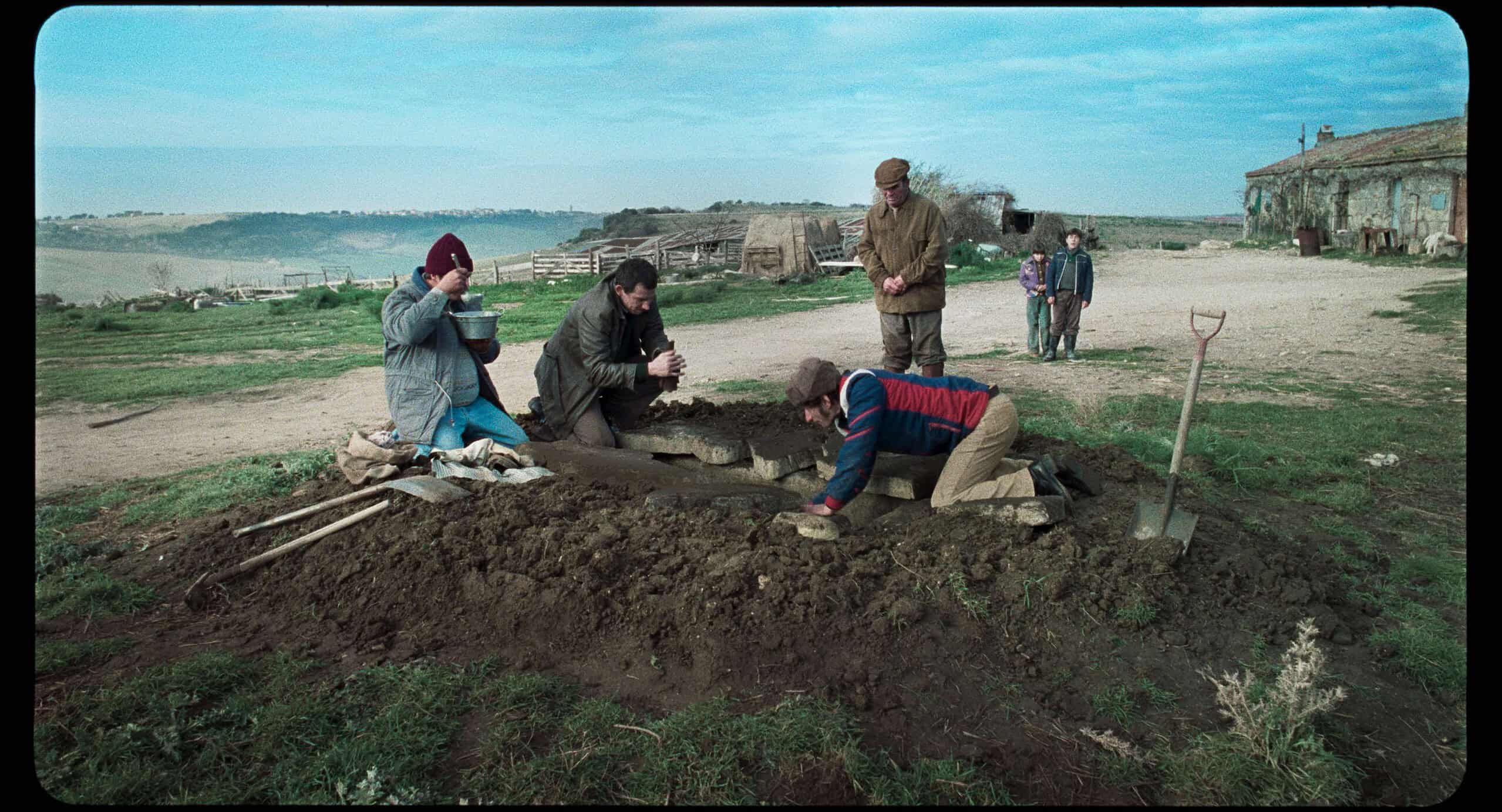 Men laboring outdoors, digging a hole in the ground near an old rural house, under a cloudy sky.