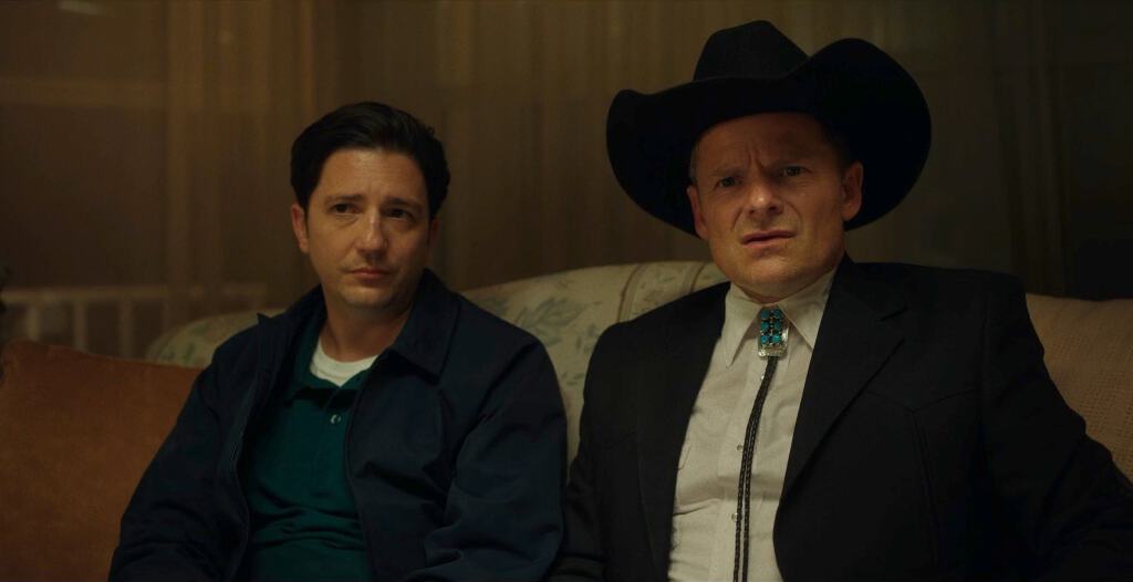 Two men on a couch with contrasting attire: one casual, one in a cowboy hat and formal wear.