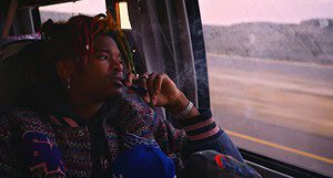 A man with colorful dreadlocks looks thoughtfully out the window of a moving bus, holding a smartphone to his ear.