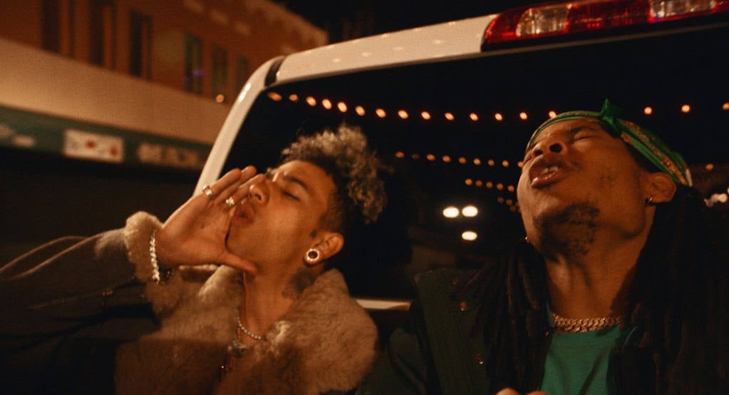 Two young men in streetwear stand next to a white van at night, one blowing a kiss and the other looking away with a relaxed expression.