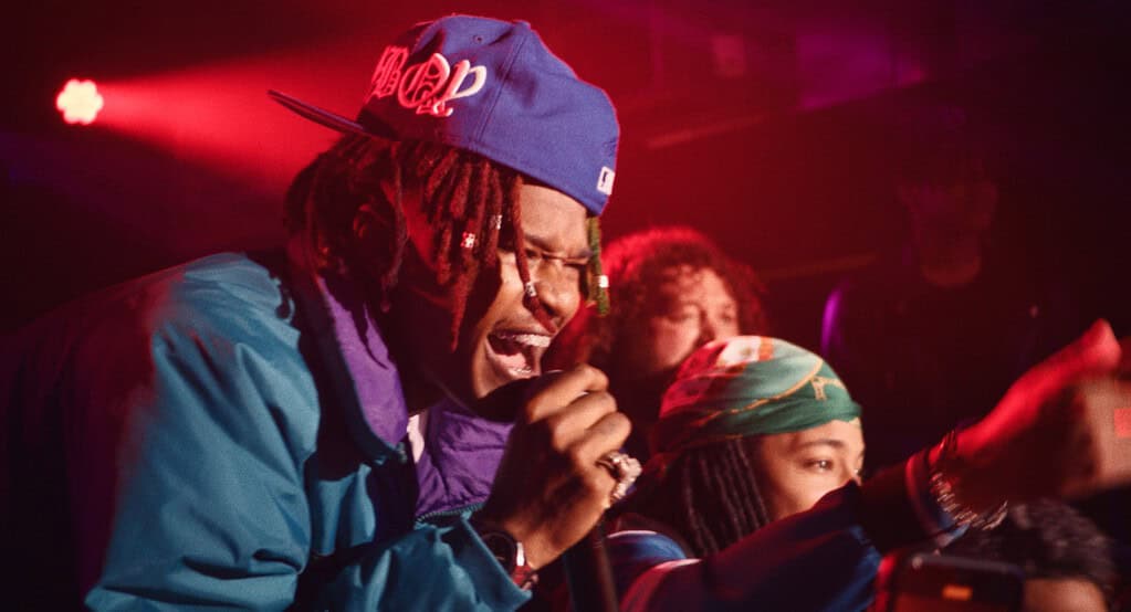 A male rapper performing on stage with a microphone, wearing a blue cap and jacket, dramatically illuminated by red stage lights.