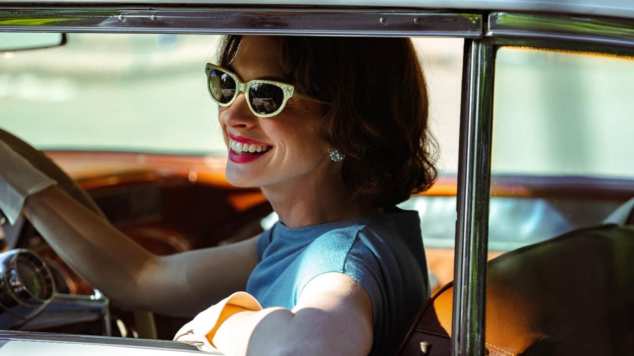 A woman in sunglasses smiling while seated at the wheel of a vintage car.