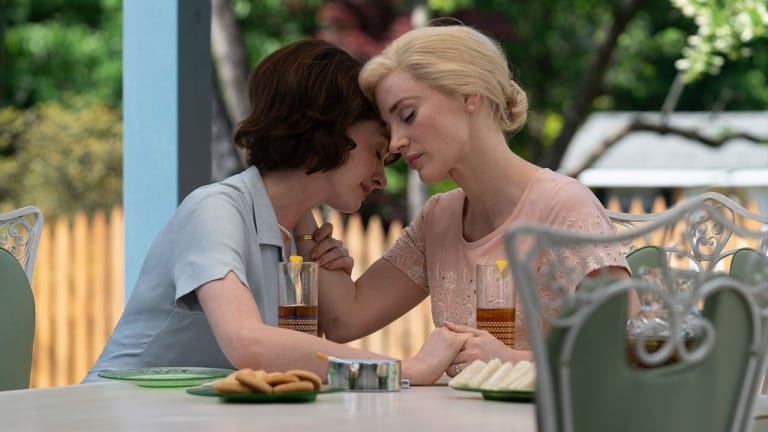 Two women sitting face to face at a table, leaning in closely, with a sense of intimacy in their interaction.